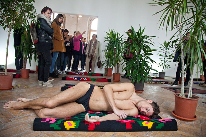 Fotografija Borisa Burića prikazuje muškarca, učesnika u radu Ivane Ivković “Spavaonica”, koji obnažen leži na ćilimu a iznad njega je publika.